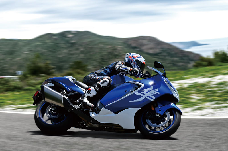 Load image into Gallery viewer, A blue and white 2026 Suzuki Hayabusa GSX 1300 RR Special Edition supersport motorcycle with a rider in full racing gear, captured in motion on a paved road with green hills and a cloudy sky in the background.