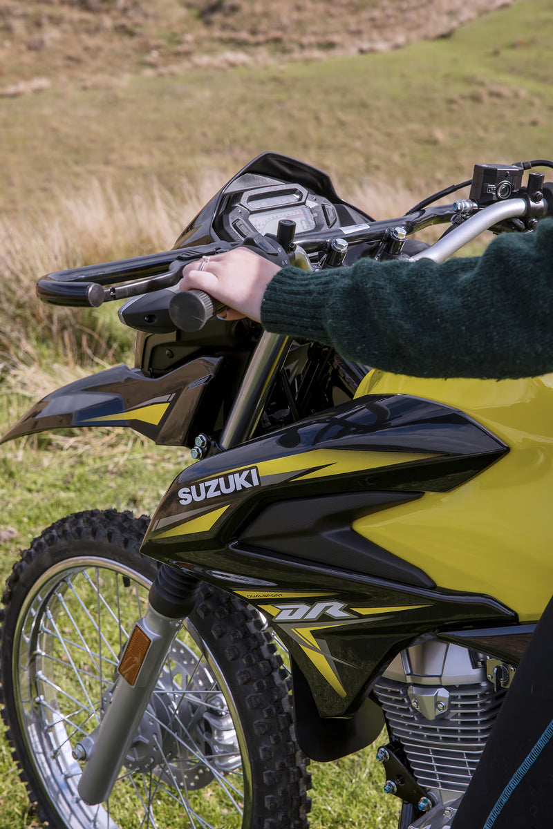 Load image into Gallery viewer, Close-up of a yellow and black 2026 Suzuki DR150 dirt bike outdoors, with a person's hand resting on the handlebar in a grassy field.