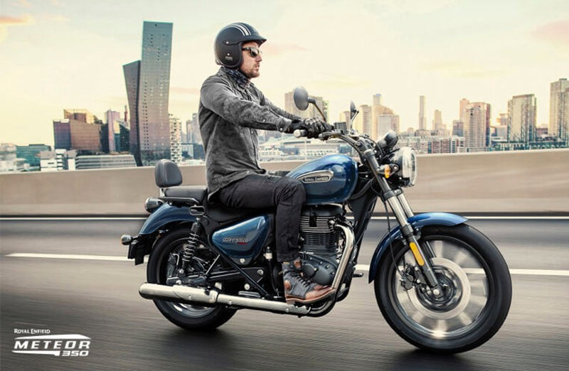 Load image into Gallery viewer, A man wearing a black helmet rides a Royal Enfield Meteor 350 motorcycle on a highway with a blurred city skyline in the background. The motorcycle is dark blue with chrome accents.