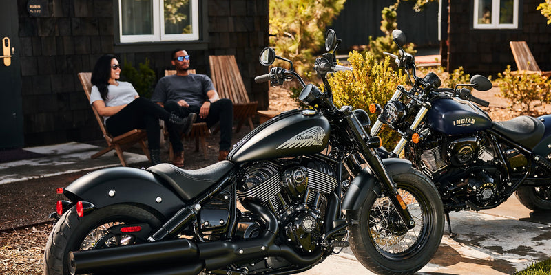 Load image into Gallery viewer, Two motorcycles parked outdoors with people sitting in the background.