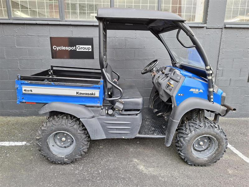 Load image into Gallery viewer, A blue and gray Kawasaki 4x4 utility vehicle parked outdoors in front of a gray brick building. The vehicle features a black roll cage, a black cargo bed, and a blue cab with visible Kawasaki branding.