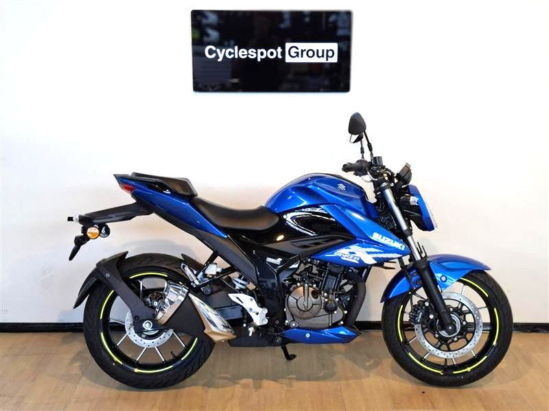 Load image into Gallery viewer, A blue and black Suzuki motorcycle displayed indoors against a white wall, with a visible 'Cyclespot Group' sign above it and a wood floor beneath.