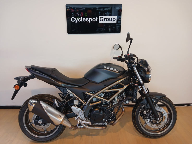 Load image into Gallery viewer, A black 2026 Suzuki SV 650 ABS motorcycle with a treble-cradle frame and chrome exhaust, parked indoors against a beige wall with a 'Cyclespot Group' sign above it.