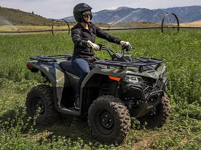 Load image into Gallery viewer, A woman wearing a black helmet and riding gear sits on a green CFMTOTO CFORCE 400 all-terrain vehicle (ATV) in a green field with mountains in the background.