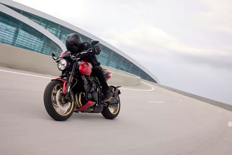 Load image into Gallery viewer, A red and black Triumph Trident motorcycle with gold accents is ridden on a curved road with a modern glass building in the background. The rider is wearing a black helmet and black riding gear.