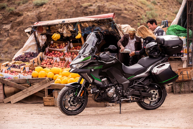 Load image into Gallery viewer, A black and green 2024 Kawasaki Versys 1000 S motorcycle with hard pannier bags is parked at a dusty outdoor market stall with people and fruit displays in the background.
