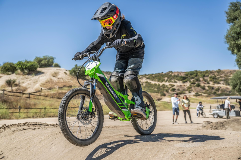 Load image into Gallery viewer, Child riding a green Kawasaki electric Elektrode bike on a dirt track with a clear blue sky and trees in the background.