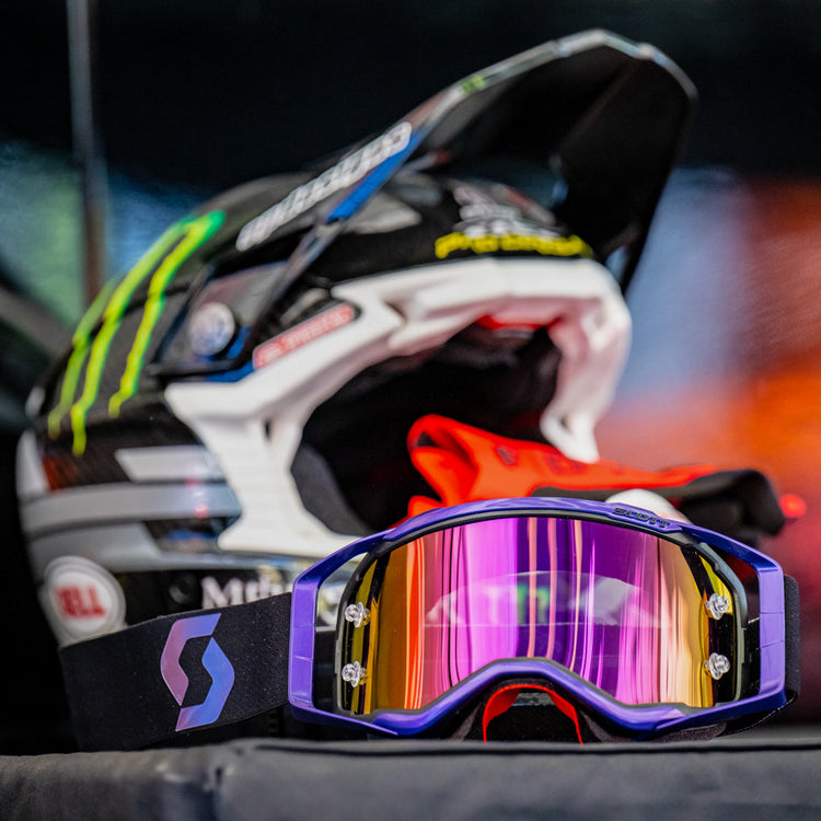 A pair of Scott motocross goggles with a purple frame and iridescent mirror lens, displayed with a Bell MX helmet. Essential eyewear for impact protection and clarity. Available at Cyclespot Auckland, Taranaki, Wellington, and online.