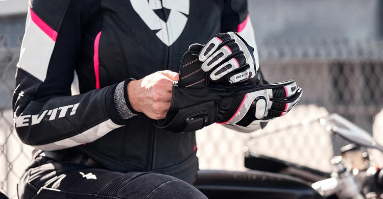 A female rider wearing a black and white REV'IT! leather motorcycle jacket with pink accents, adjusting her matching gauntlet gloves while seated on a bike.