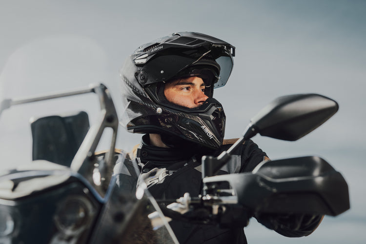 An adventure motorcycle rider wearing a black Airoh Commander dual-sport helmet with the visor raised. This carbon fibre ADV helmet is ideal for touring and offroad adventures, available at Cyclespot Auckland, Taranaki, and Wellington.