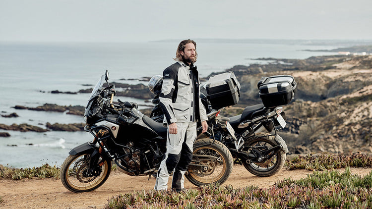 A male motorcyclist standing next to a loaded adventure bike on a coastal cliffside, wearing a grey and black Richa technical touring jacket and matching trousers.