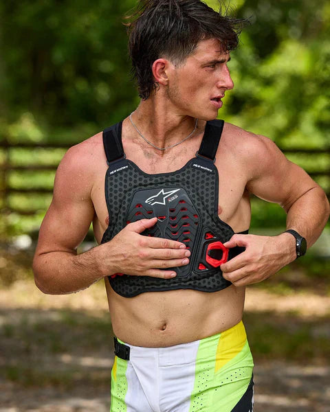 Motocross rider outdoors wearing an Alpinestars chest protector over his bare torso. The black and red roost guard features a honeycomb ventilation design and prominent logo. The rider adjusts a side buckle, paired with white and yellow racing pants.
