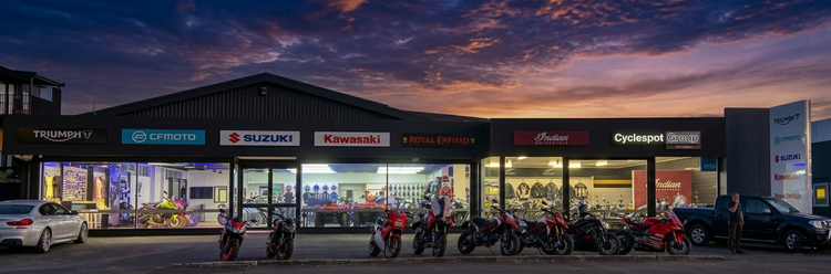 Panoramic sunrise view of the Cyclespot Taranaki motorcycle store, featuring a professionally lit showroom with Triumph, Suzuki, Kawasaki, Indian, CFMoto and Royal Enfield branding and a lineup of motorcycles out front.