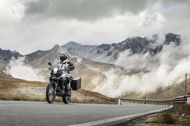 An adventure motorcycle rider in full technical wet weather gear navigating a winding mountain road through clouds and mist, demonstrating high-performance touring apparel available at Cyclespot NZ.
