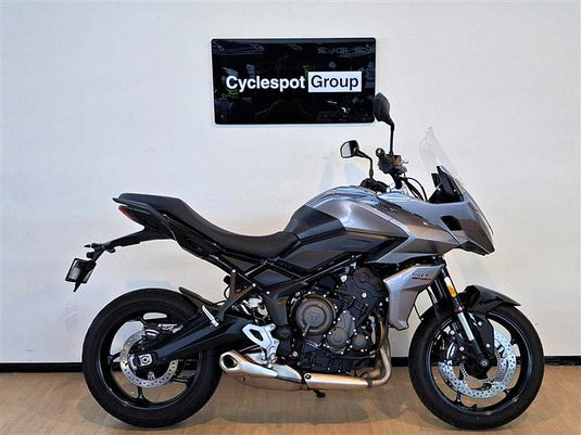 A silver and black  2023 Triumph Tiger Sport 660 motorcycle is parked indoors against a white wall with a 'Cyclespot Group' sign above it. The bike is shown in full side profile with its front wheel turned slightly inward.