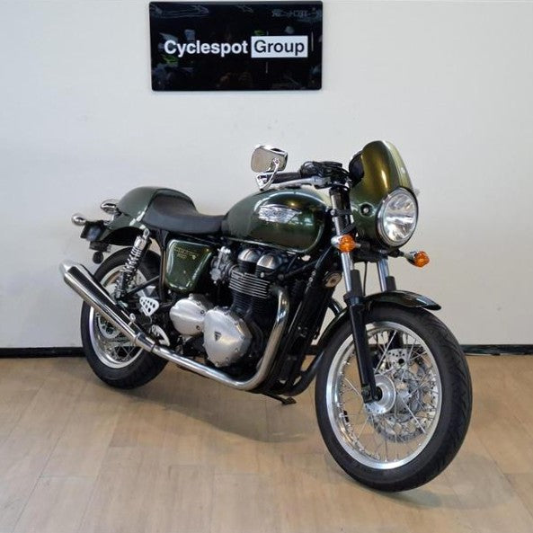 Load image into Gallery viewer, A classic-style 2013 Triumph Thruxton 900 motorcycle with a dark green metallic finish and chrome engine components, parked indoors on a light wood floor against a white wall with a 'Cyclespot Group' sign above it.
