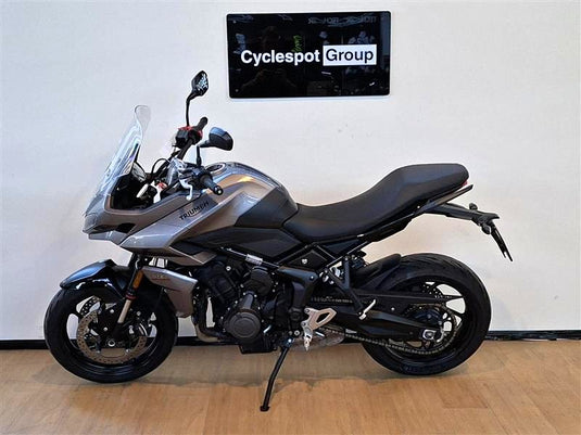 A silver and black  2023 Triumph Tiger Sport 660 Adventure motorcycle is parked indoors on a wooden floor against a white wall, with a 'Cyclespot Group' sign visible on the wall behind it.