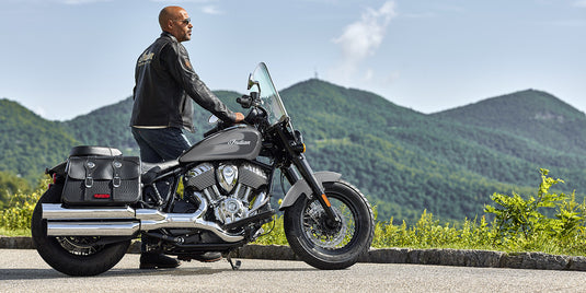 A man wearing a black leather jacket and jeans sits on a black and silver Indian Super Chief motorcycle with saddlebags, parked outdoors on a paved road with green mountains in the background.