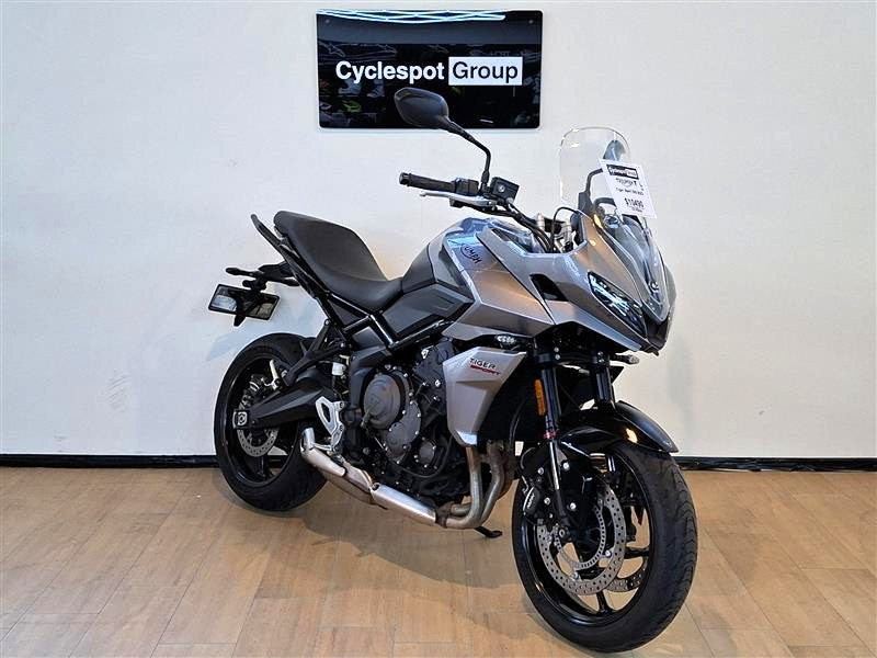 Load image into Gallery viewer, A silver and black  2023 Triumph Tiger Sport 660 motorcycle is parked indoors on a light wood floor against a white wall, with a 'Cyclespot Group' sign visible above it.
