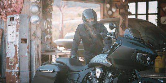 A person in a black helmet and dark jacket stands beside a large black 2026 Indian Challenger Limited in a rustic, sunlit garage or workshop with vintage equipment and wooden posts.