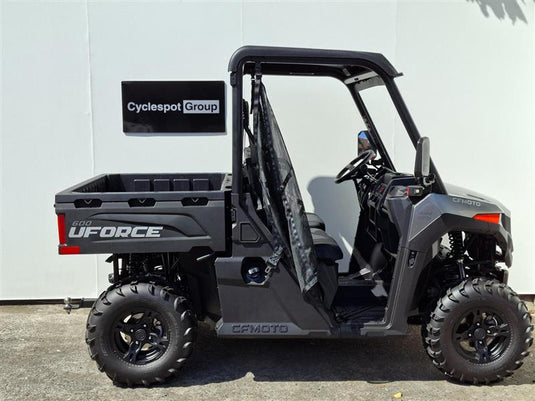 A black and gray 2026 CFMOTO UFORCE 600 EPS side-by-side utility vehicle with an open side door and roll cage, parked against a white wall. A black sign reading 'Cyclespot Group' is visible behind the vehicle.