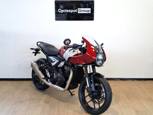 A diagonal view from the front of a 2026 Triumph Thruxton 400 in Carnival Red on a wooden floor, backed by a white wall with a Cyclespot Group sign in white on black.