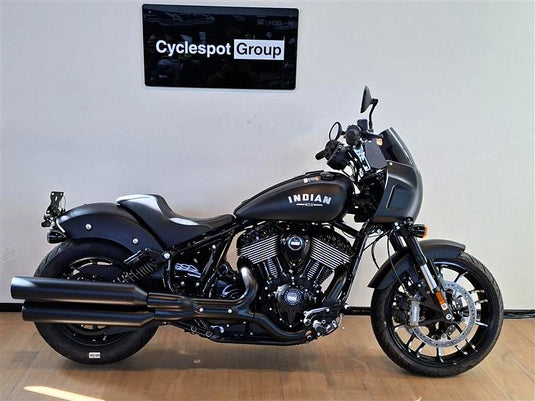 A black Indian Sport Chief cruiser motorcycle is parked indoors against a white wall, with a visible 'Cyclespot Group' sign above it. The bike has chrome engine components, black tires, and white 'INDIAN' branding on the fuel tank.