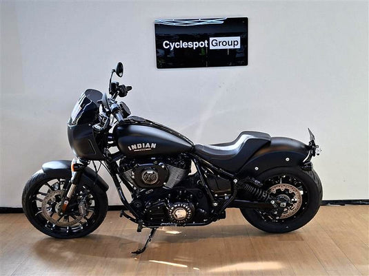 A black Indian Sport Chief motorcycle with chrome accents is parked indoors on a wooden floor against a white wall, with a sign reading 'Cyclespot Group' on the wall behind it.