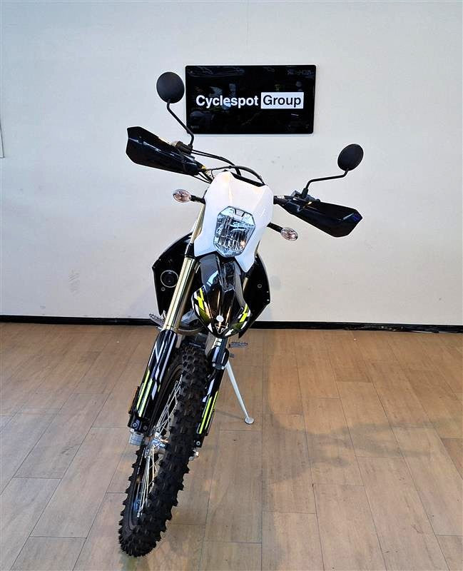 Load image into Gallery viewer, A black and white Triumph TF 250 E motorcycle with yellow accents, displayed on a kickstand in an indoor showroom with a wood floor and a wall sign reading 'Cyclespot Group'.
