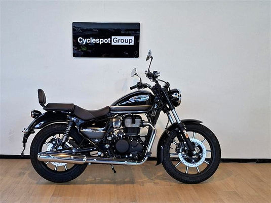 A black and silver Royal Enfield cruiser motorcycle with a brown seat, displayed in profile on a wooden floor against a light gray wall. A 'Cyclespot Group' sign is mounted on the wall above the motorcycle.