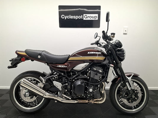 A Kawasaki Z900RS motorcycle in a dark maroon and black color scheme with gold stripes, displayed indoors against a white wall with a 'Cyclespot Group' sign above it.