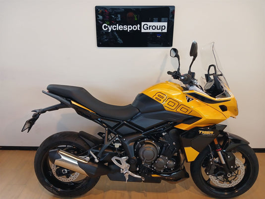 A yellow and black Triumph Tiger 800motorcycle is parked indoors against a light beige wall, with a 'Cyclespot Group' sign visible above it.