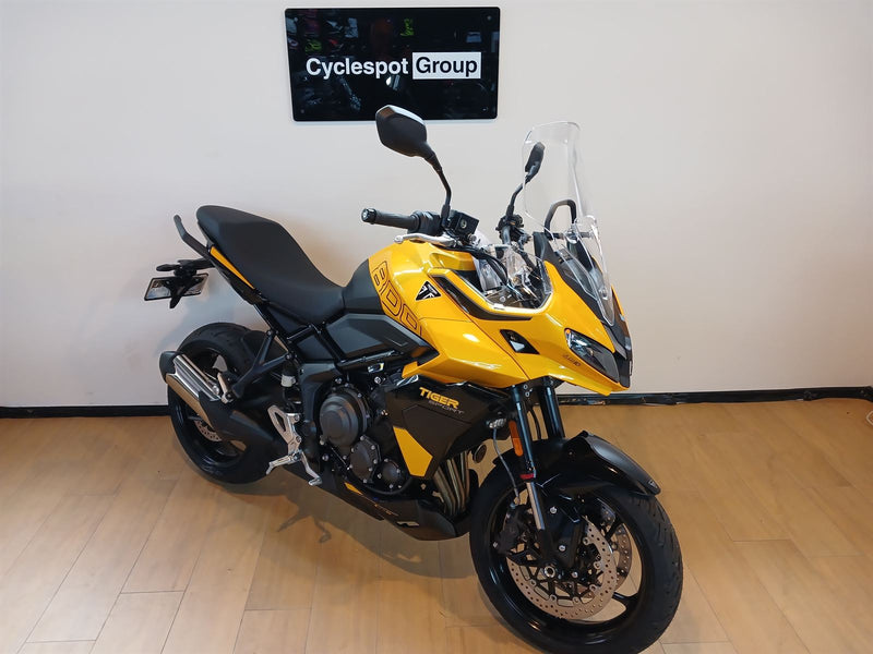 Load image into Gallery viewer, A yellow and black Triumph Tiger 800 motorcycle with a clear windshield, parked indoors on a light wood floor against a white wall with a 'Cyclespot Group' sign above it.
