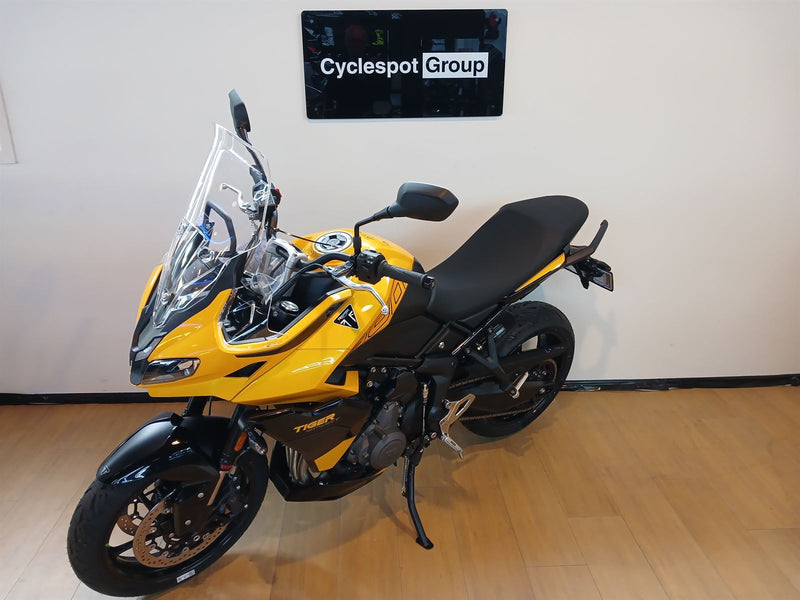 Load image into Gallery viewer, A yellow and black Triumph Tiger 800 motorcycle with a tall windscreen is parked indoors on a wooden floor against a white wall, with a sign reading 'Cyclespot Group' mounted above it.
