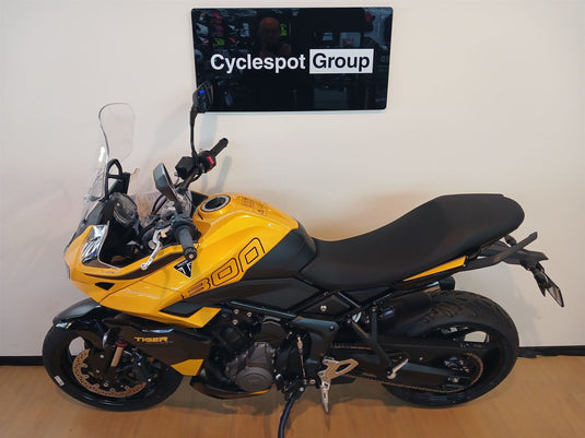 A yellow and black Triumph Tiger 800 motorcycle parked indoors against a beige wall, with a 'Cyclespot Group' sign visible above it.