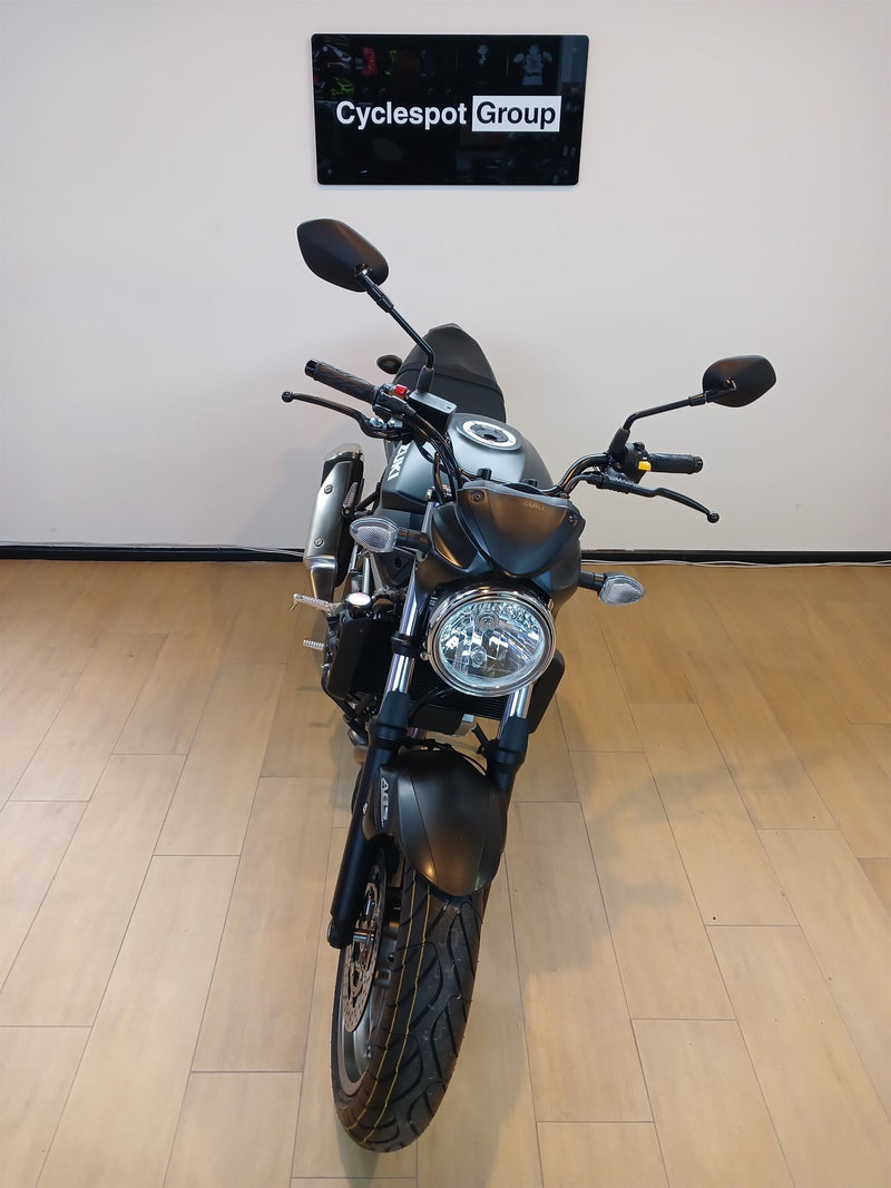 Load image into Gallery viewer, A black and silver cruiser motorcycle is photographed from the front inside a showroom with light wood flooring and a white wall. A sign on the wall reads 'Cyclespot Group'.
