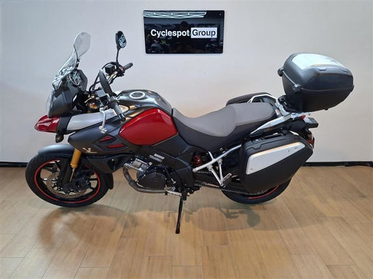 A red and black Suzuki DL1000 V-Strom adventure-touring motorcycle with gray saddlebags and a black top case, parked indoors on a wood floor against a light gray wall with a 'Cyclespot Group' sign.