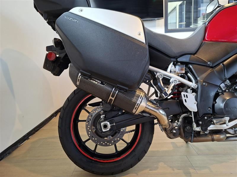 Load image into Gallery viewer, Close-up of the rear section of a Suzuki DL1000 V-Strom motorcycle, showing the black and silver hard saddlebag, carbon-fiber-look exhaust, rear wheel with a red rim, and part of the red seat on a showroom floor.
