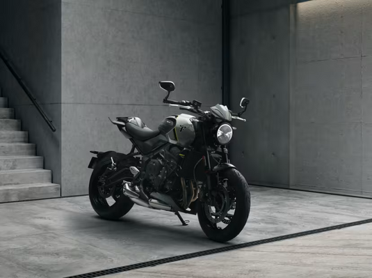 A black and silver 2026 Triumph Trident 660 LAMS motorcycle with yellow accents is parked on a concrete floor in a minimalist industrial setting with concrete walls and stairs.