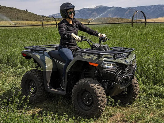 A woman wearing a black helmet and riding gear sits on a 2025 CFMoto CFORCE 400 Hunter Green all-terrain vehicle (ATV) in a green field with mountains in the background.