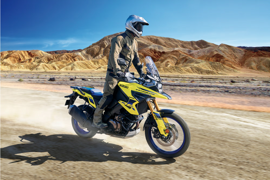A rider in full off-road gear sits on a yellow and black 2025 Suzuki V-Strom 1050 RJ adventure motorcycle, kicking up dust on a desert road with rocky mountains and a blue sky in the background.