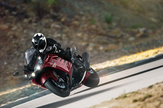 A red and black 2025 Kawasaki Ninja ZX 14 R SE motorcycle with a rider in black gear and a white helmet, leaning sharply during a turn on a paved road with a blurred natural background.