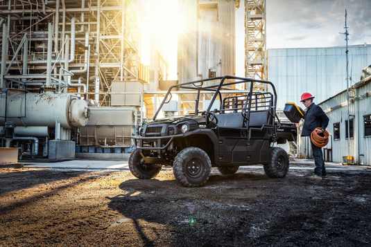 An all-terrain vehicle is parked on a dirt lot next to a white industrial building, with a man in a red hard hat standing nearby holding a yellow item. The scene is backlit by bright sunlight, casting strong shadows.