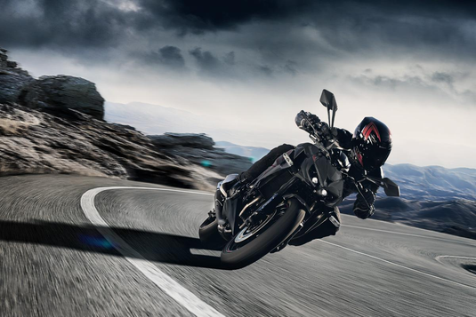 A green and black Kawasaki Z1000 motorcycle with a rider in full gear leaning into a curved mountain road under a dramatic cloudy sky.