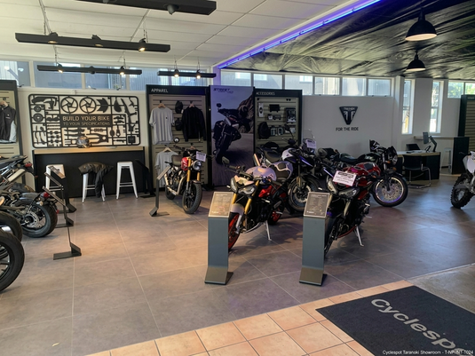 Interior view of Cyclespot Taranaki, featuring a clean, professionally lit Triumph motorcycle showroom with a dedicated service fitment area, riding apparel, and several modern Triumph bikes on display under clear geometric lighting.