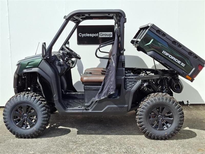 Load image into Gallery viewer, A green 2026 CFMOTO UFORCE U10 PRO side-by-side utility vehicle with a dark green front fender, brown seat, and a black cargo bed carrying a large black box, parked against a white wall.
