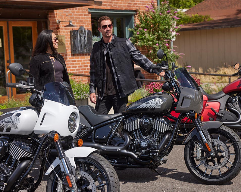 Load image into Gallery viewer, A man and a woman in black leather jackets stand outdoors beside a black Indian Sport Chief motorcycle, with other motorcycles partially visible in the foreground and a brick building in the background.

