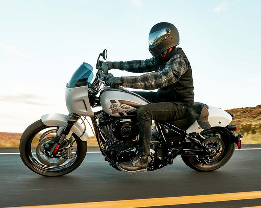 A person wearing a black helmet, plaid shirt, and black vest rides a white and black Indian Sport Chief cruiser motorcycle on a paved road with a desert landscape in the background.