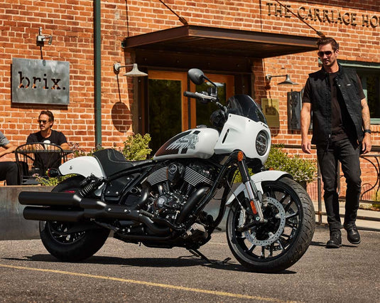 A white and black Indian Sport Chief cruiser motorcycle is parked outdoors in front of a brick building with a man walking beside it and another seated person in the background. The motorcycle has a white fuel tank and body panels with black engine, exhausts, seat, and wheels.