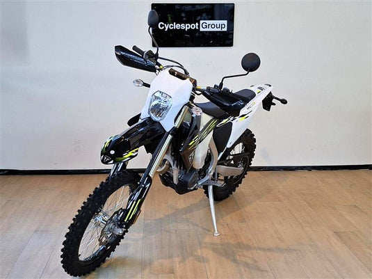 A white and black Triumph TF 450-E dirt bike with yellow accents, standing on its kickstand in an indoor showroom with a wood floor and a wall-mounted sign that reads 'Cyclespot Group'.
